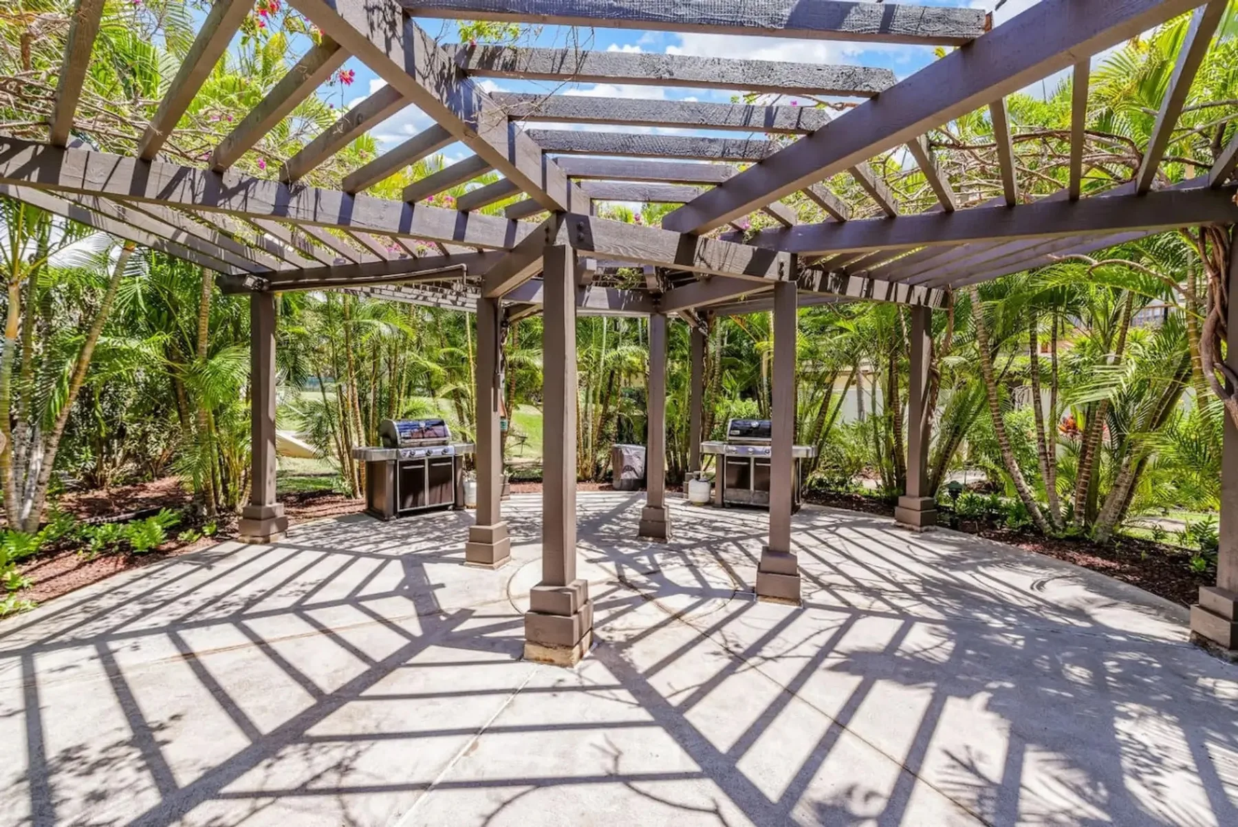 Un patio exterior pavimentado bajo una pérgola de madera, rodeado de plantas tropicales.
