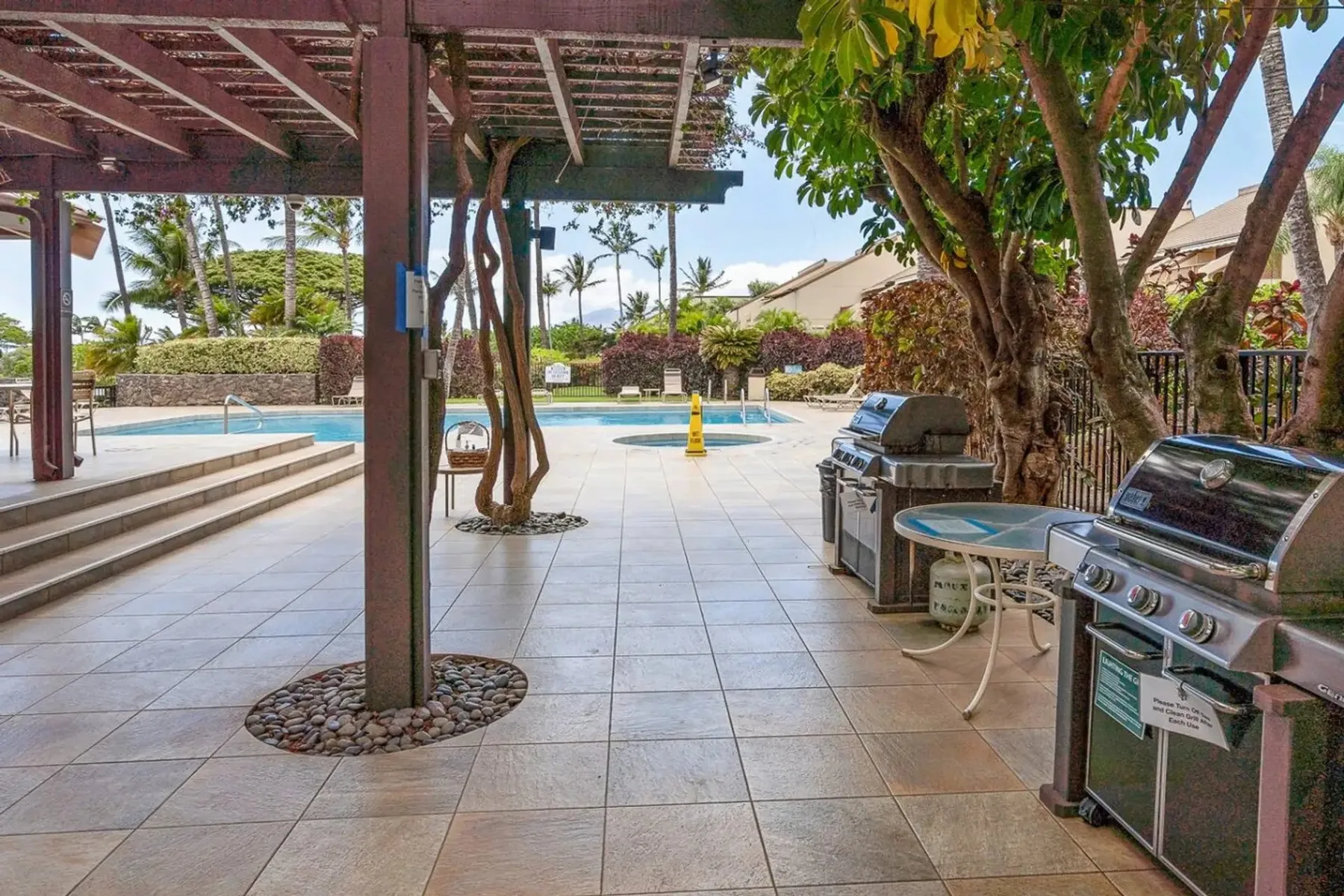 Un patio exterior embaldosado con piscina, árboles y parrillas.