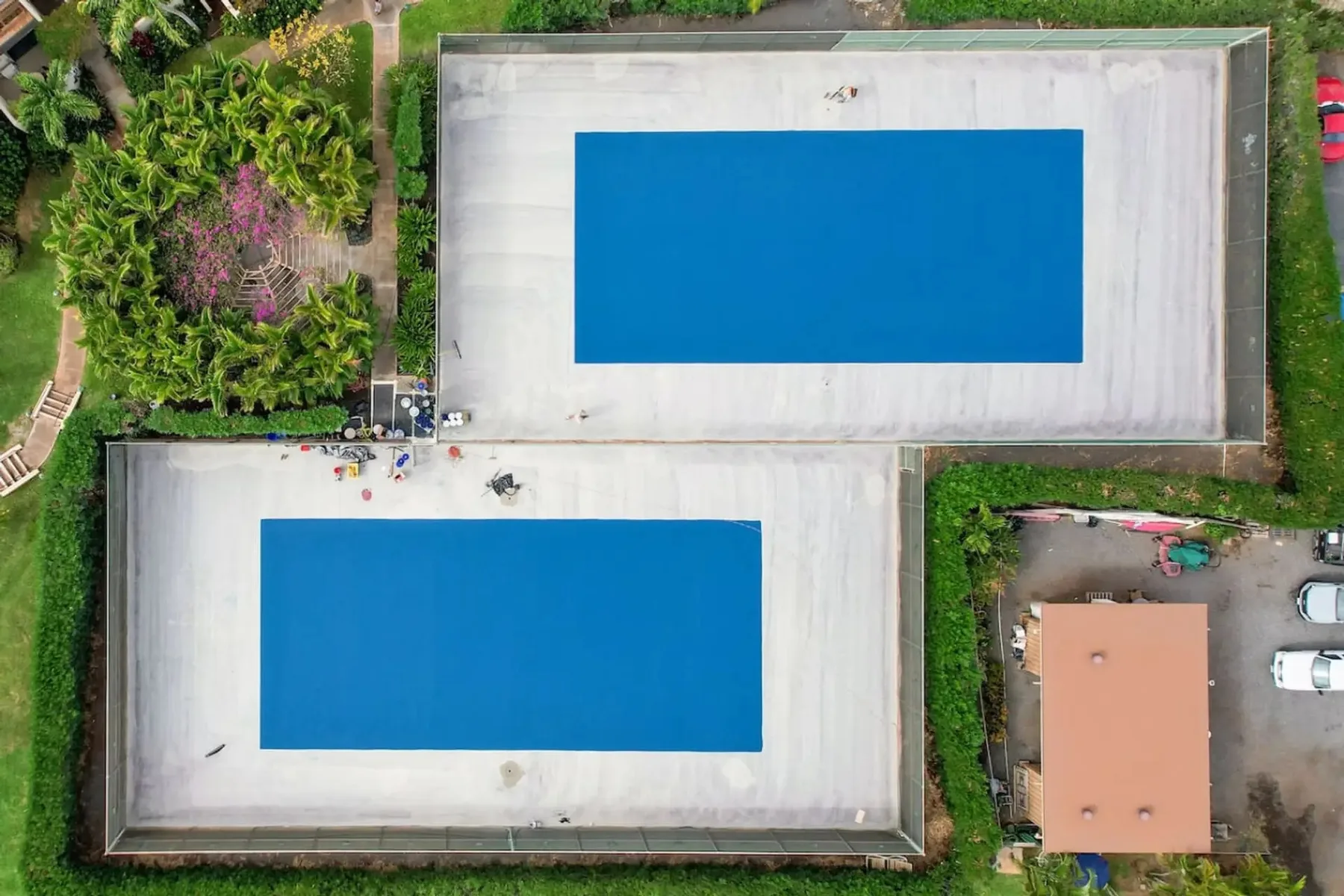 Una vista aérea muestra dos piscinas rectangulares azules rodeadas de terrazas claras y vegetación.