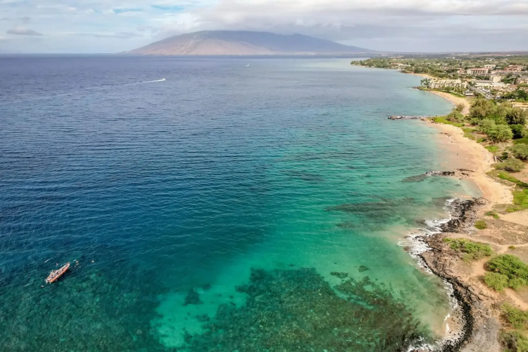 La costa se curva junto a aguas turquesas y transparentes, con colinas a lo lejos.