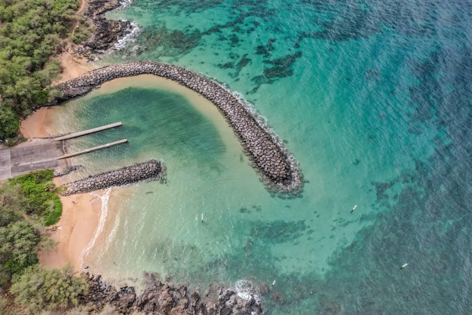 Vista aérea de una pequeña playa y un rompeolas curvo de rocas en aguas turquesas.