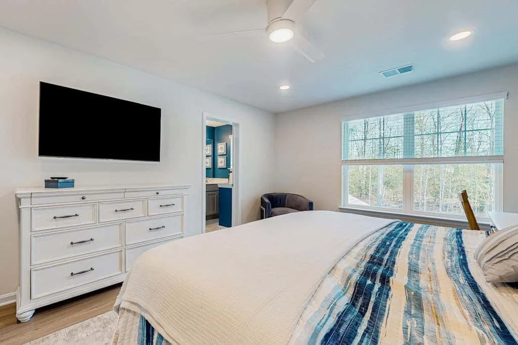 Un dormitorio luminoso con una cama, una cómoda y grandes ventanas.