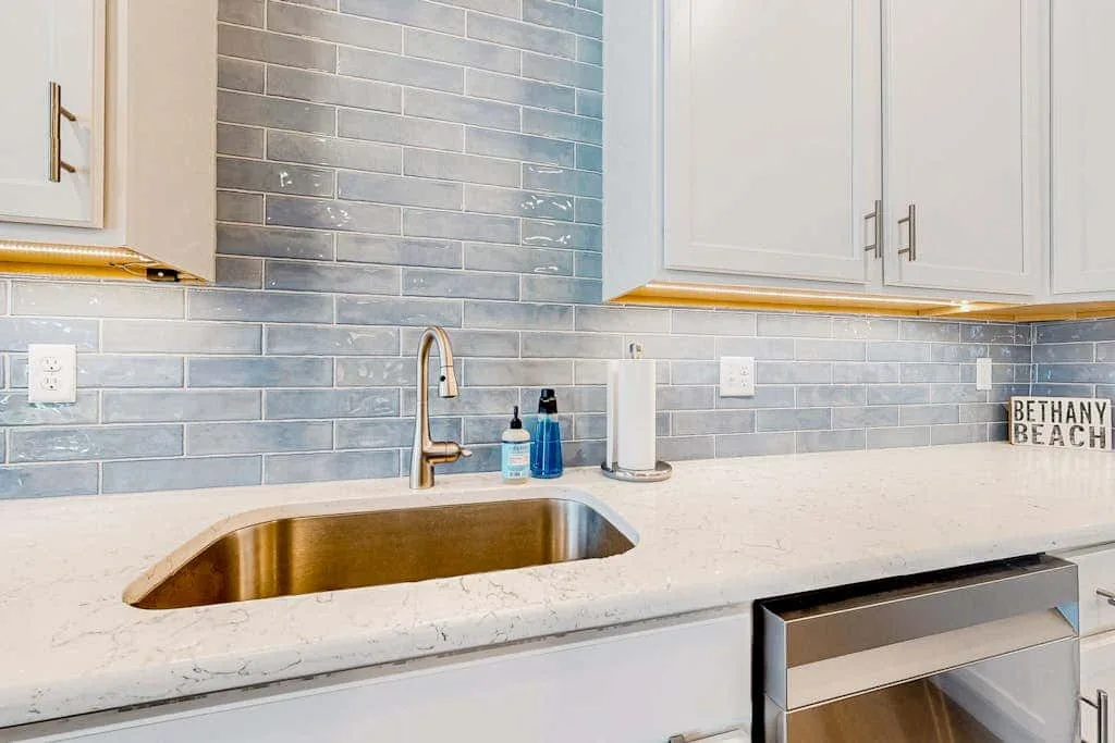 Una encimera luminosa de cocina con fregadero, grifo y salpicadero de azulejos grises.