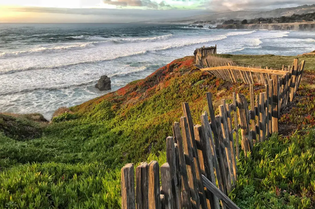 A coastal shoreline with waves washing against a grassy bluff lined by a wooden fence.