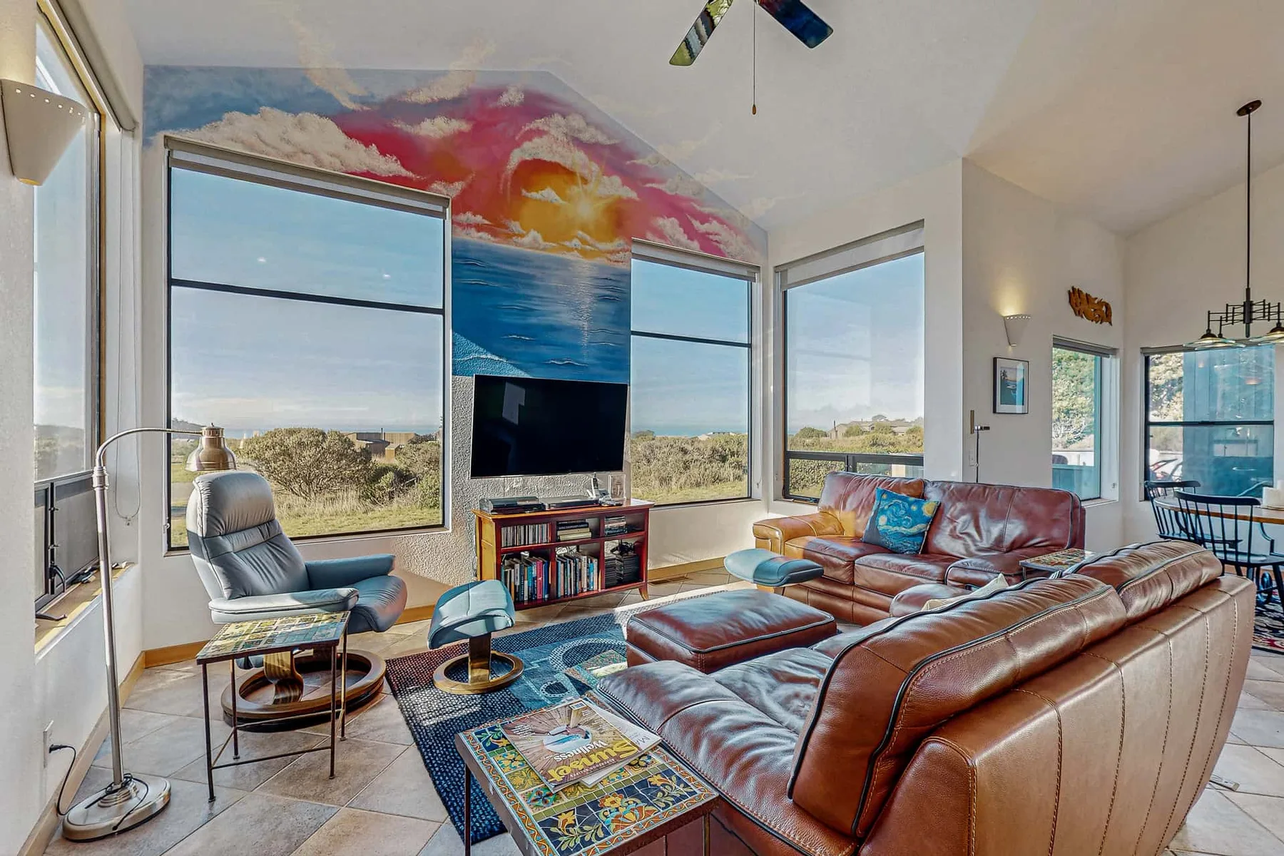 A bright living room has leather seating, large windows, and a colorful ocean mural.