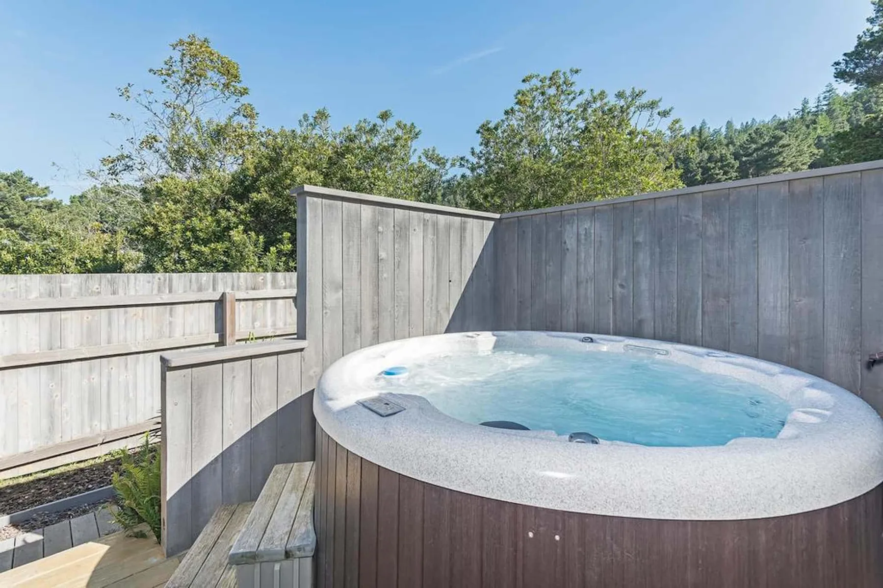 An outdoor hot tub sits within a wooden privacy fence, surrounded by trees.