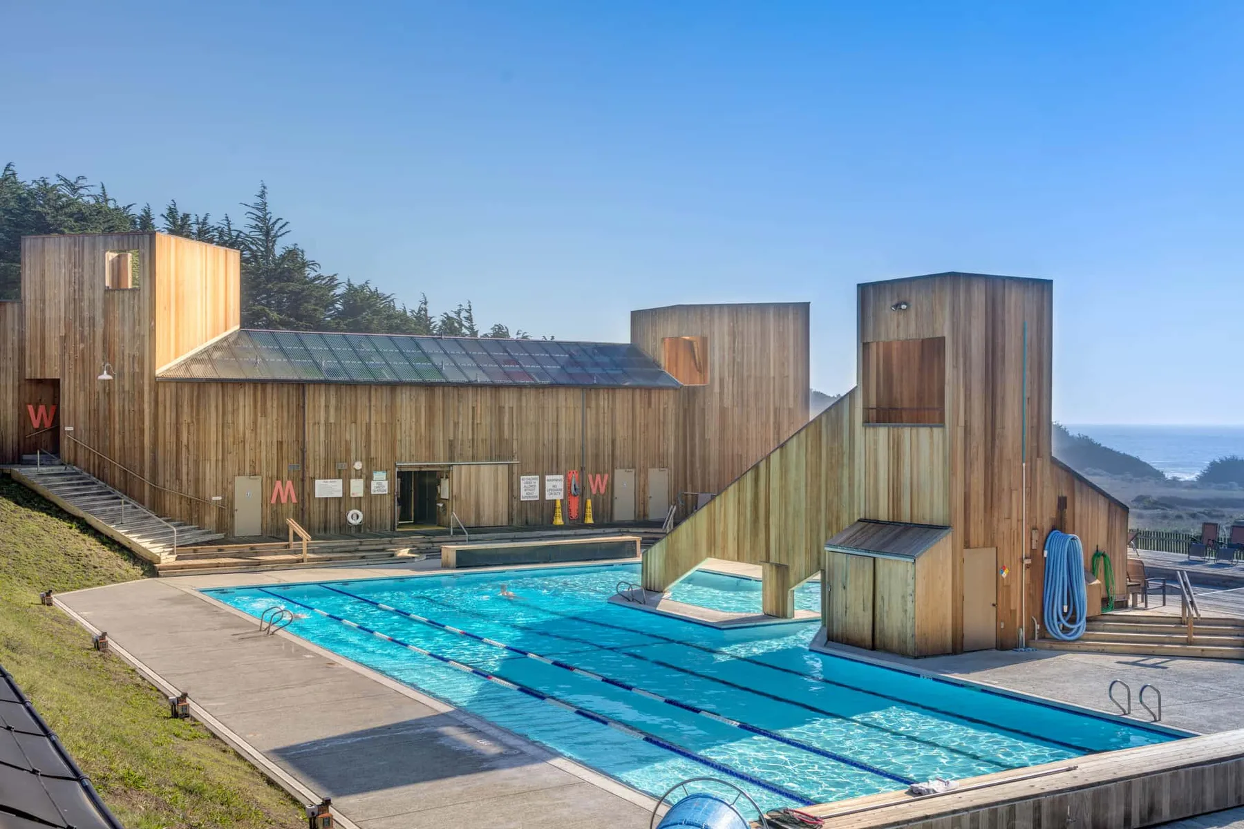An outdoor swimming pool sits beside a timber-clad building, with the sea beyond.