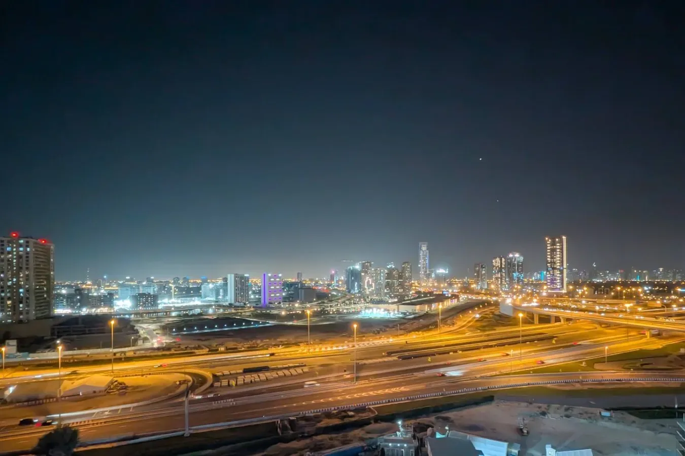 A nighttime city skyline rises beyond illuminated highways and traffic.