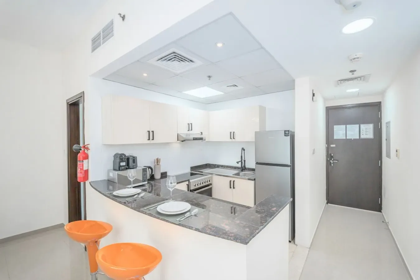 A bright kitchenette with a counter, orange stools, and a refrigerator.