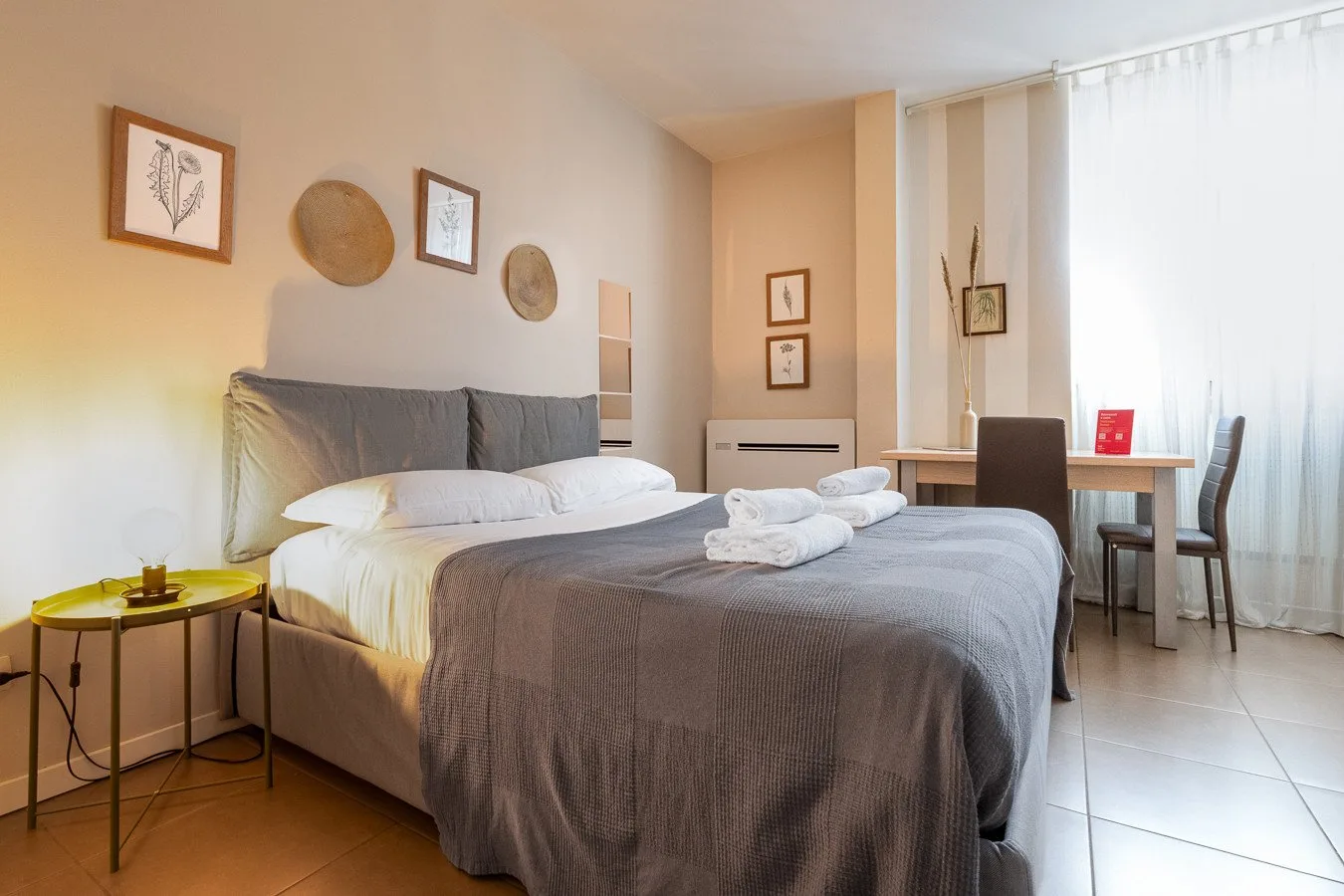 Un dormitorio con una cama bien tendida y una mesita junto a la ventana.