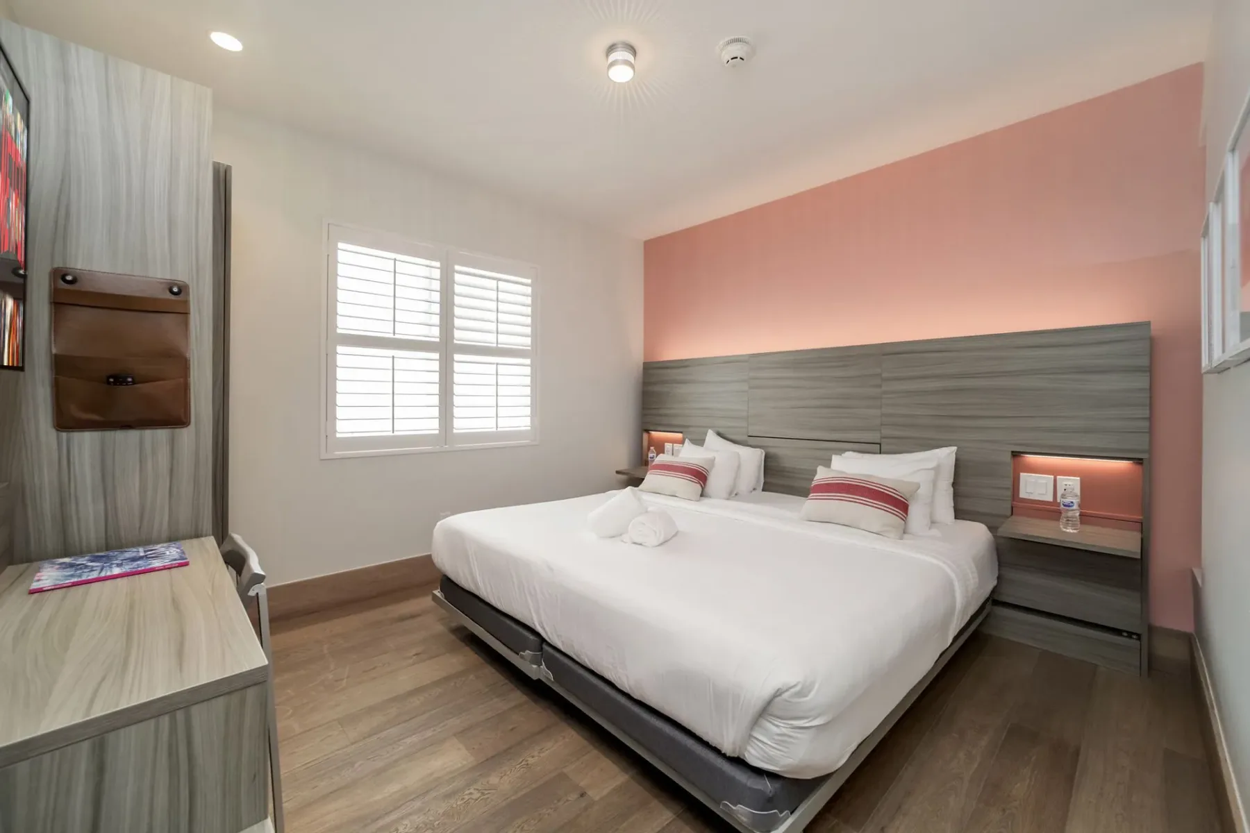 Hotel bedroom with a bed, nightstands, desk, and window; the wall behind the bed is pink.