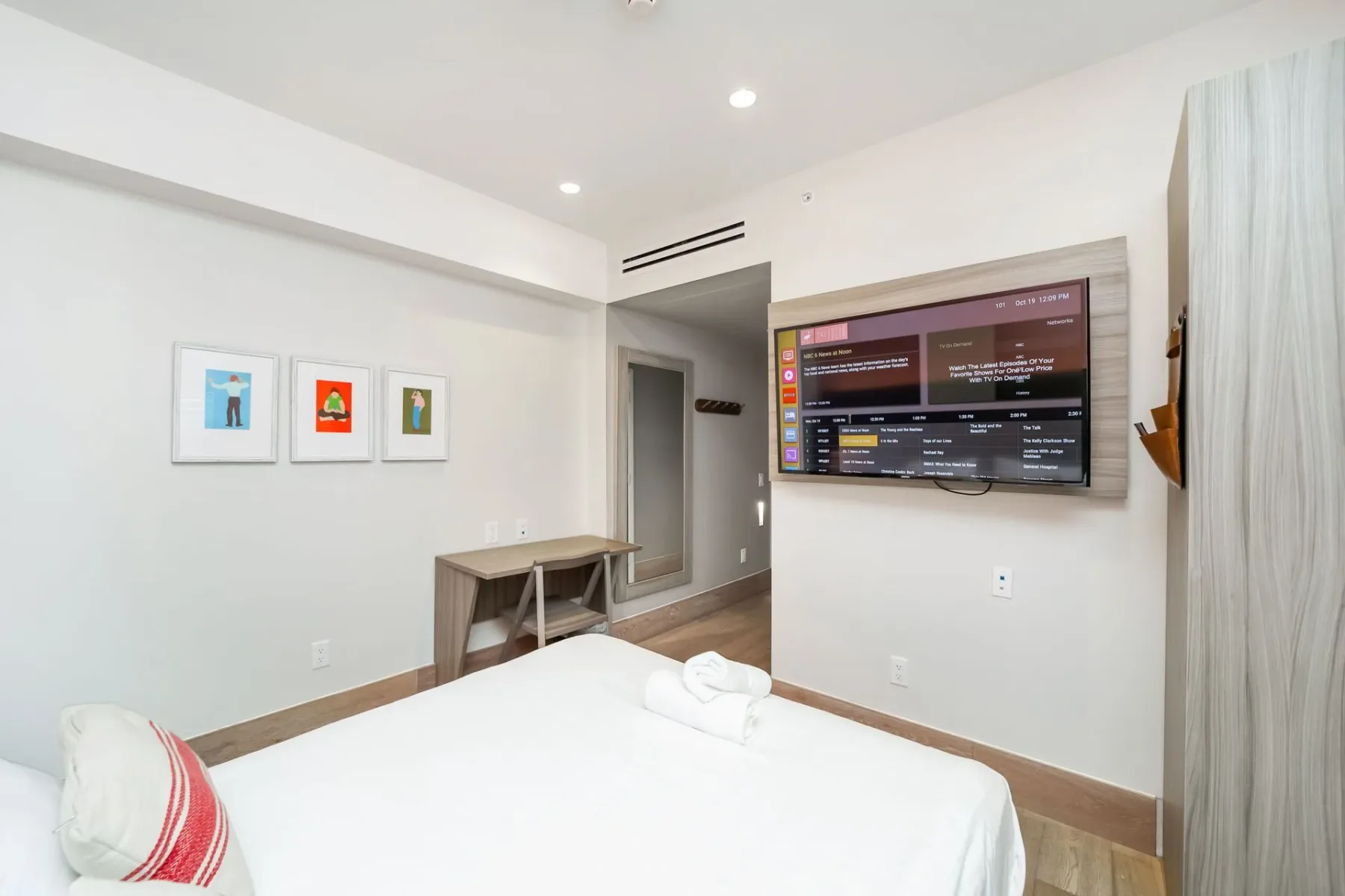 Petite bedroom with a bed, TV, artwork, and doorway to a hallway.