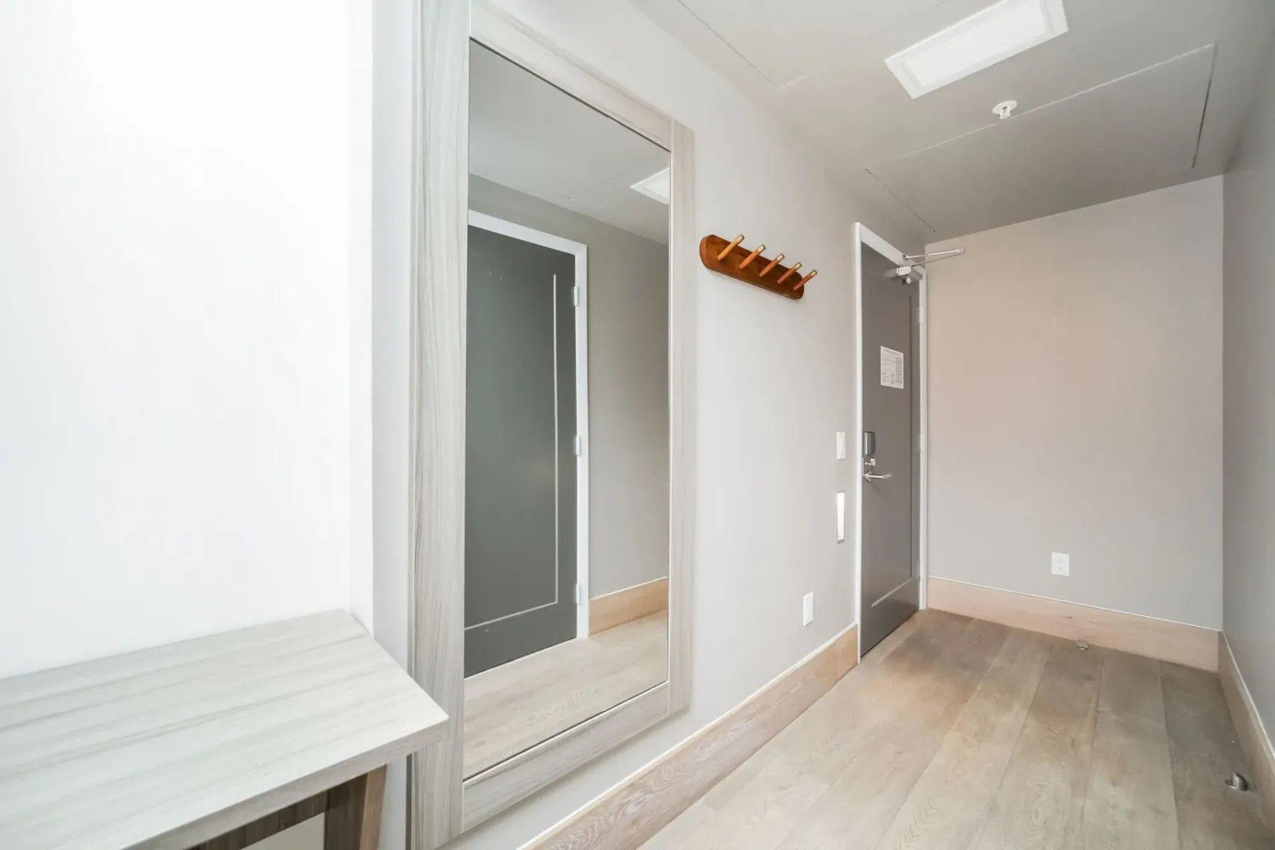 Hallway with a full-length mirror, light wood floors, gray walls, a bench, and coat rack.