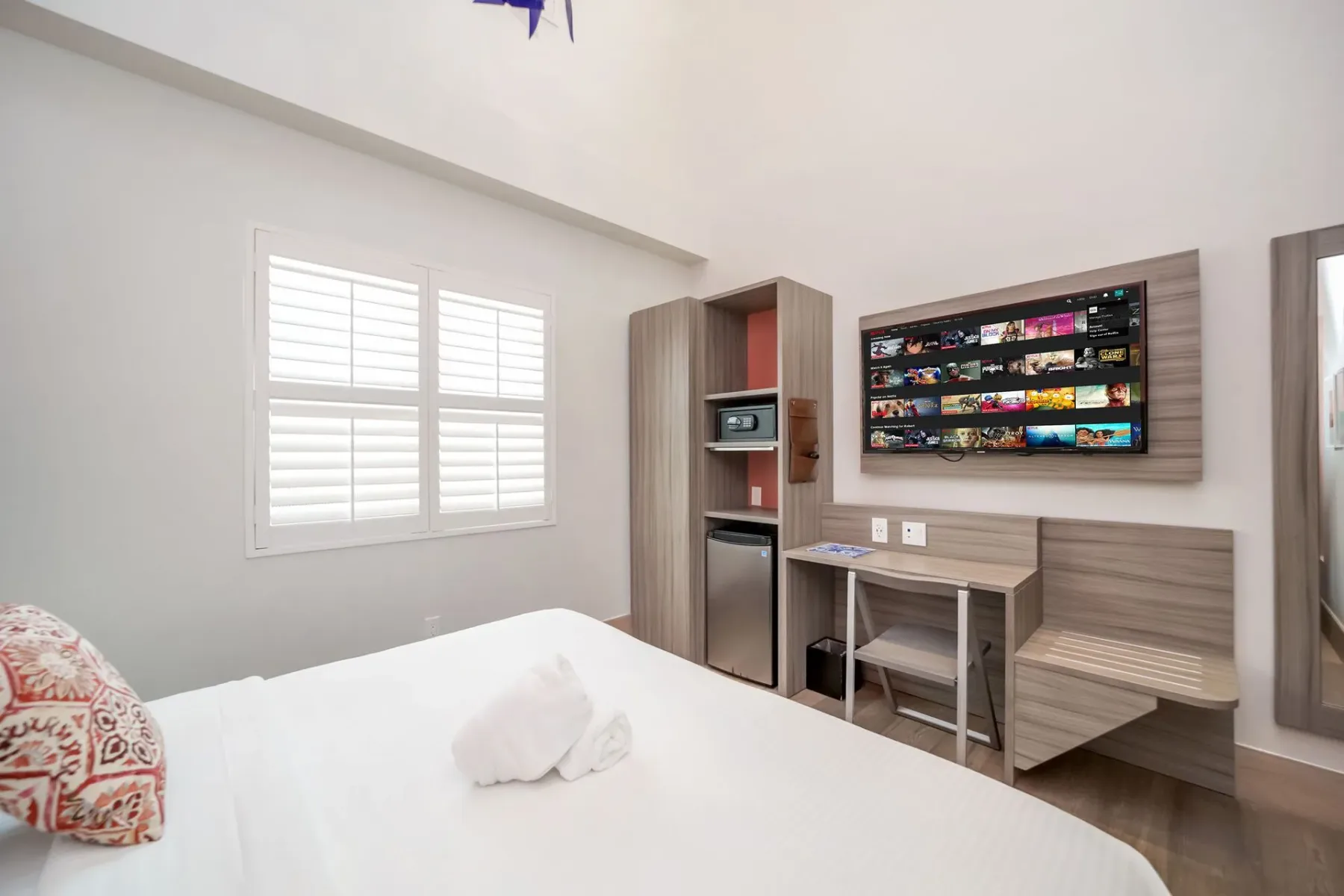 Double Height King Suite bedroom with white bedding, a TV, window with blinds, desk, and small fridge.