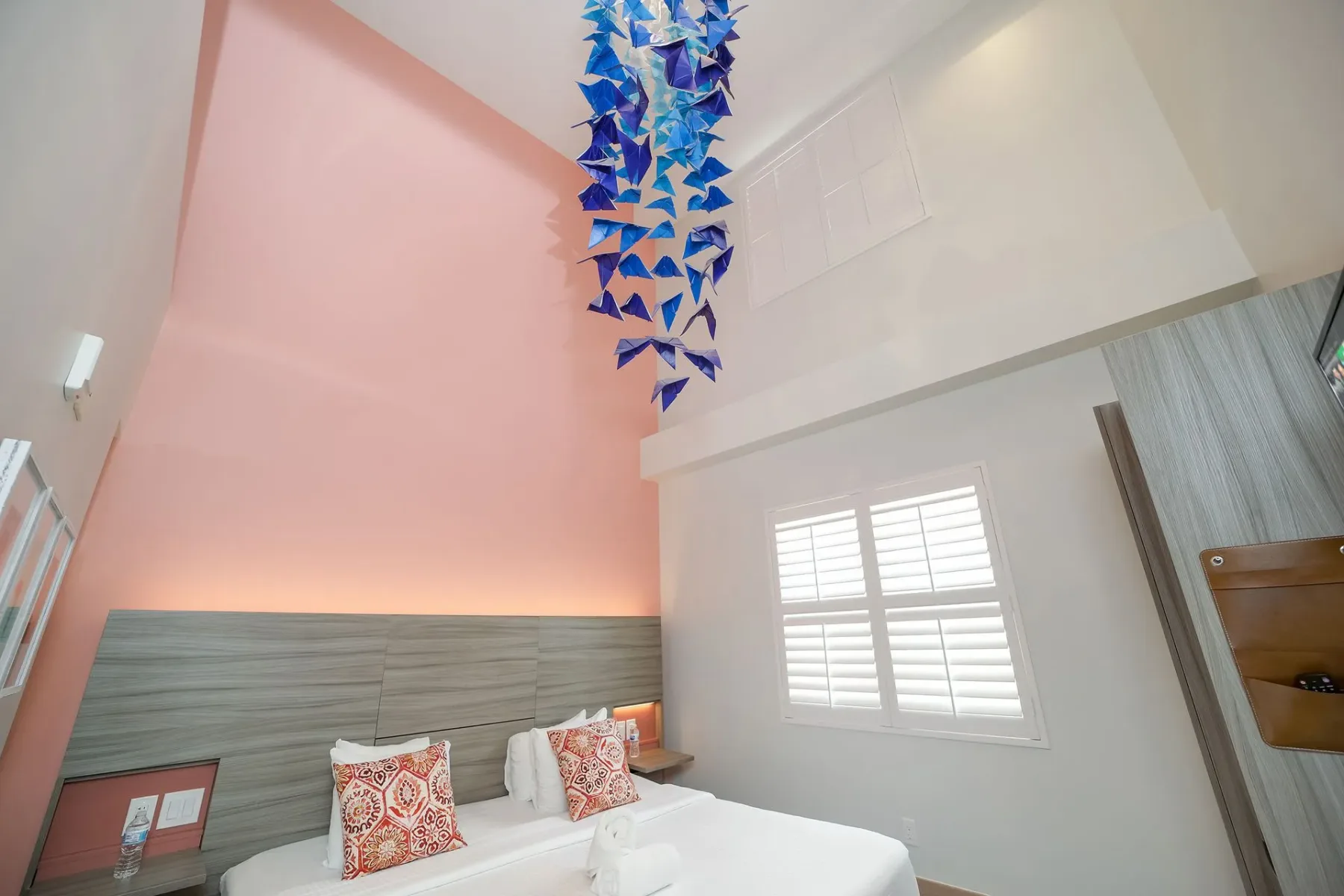 Double Height King Suite bedroom with a bed, pink and white walls, a window with shutters, and blue hanging art.