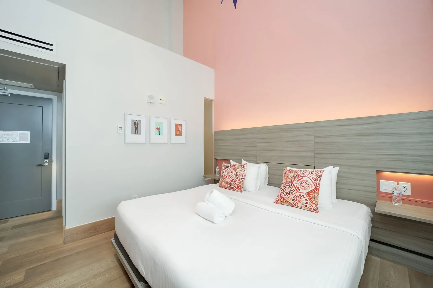 Hotel bedroom with a king-sized bed, decorative pillows, light wood floors, and a pink accent wall.
