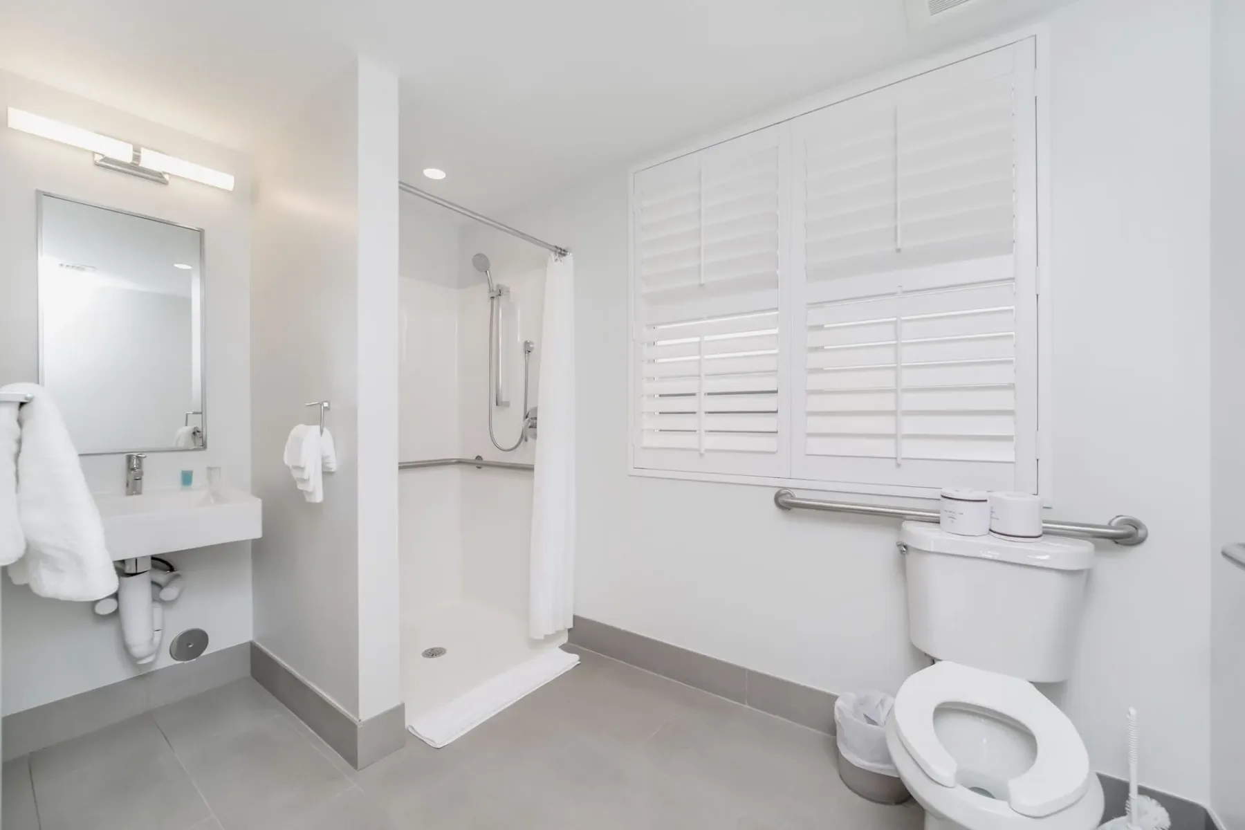 Petite bathroom with a walk-in shower, toilet with grab bar, sink, and shuttered window.