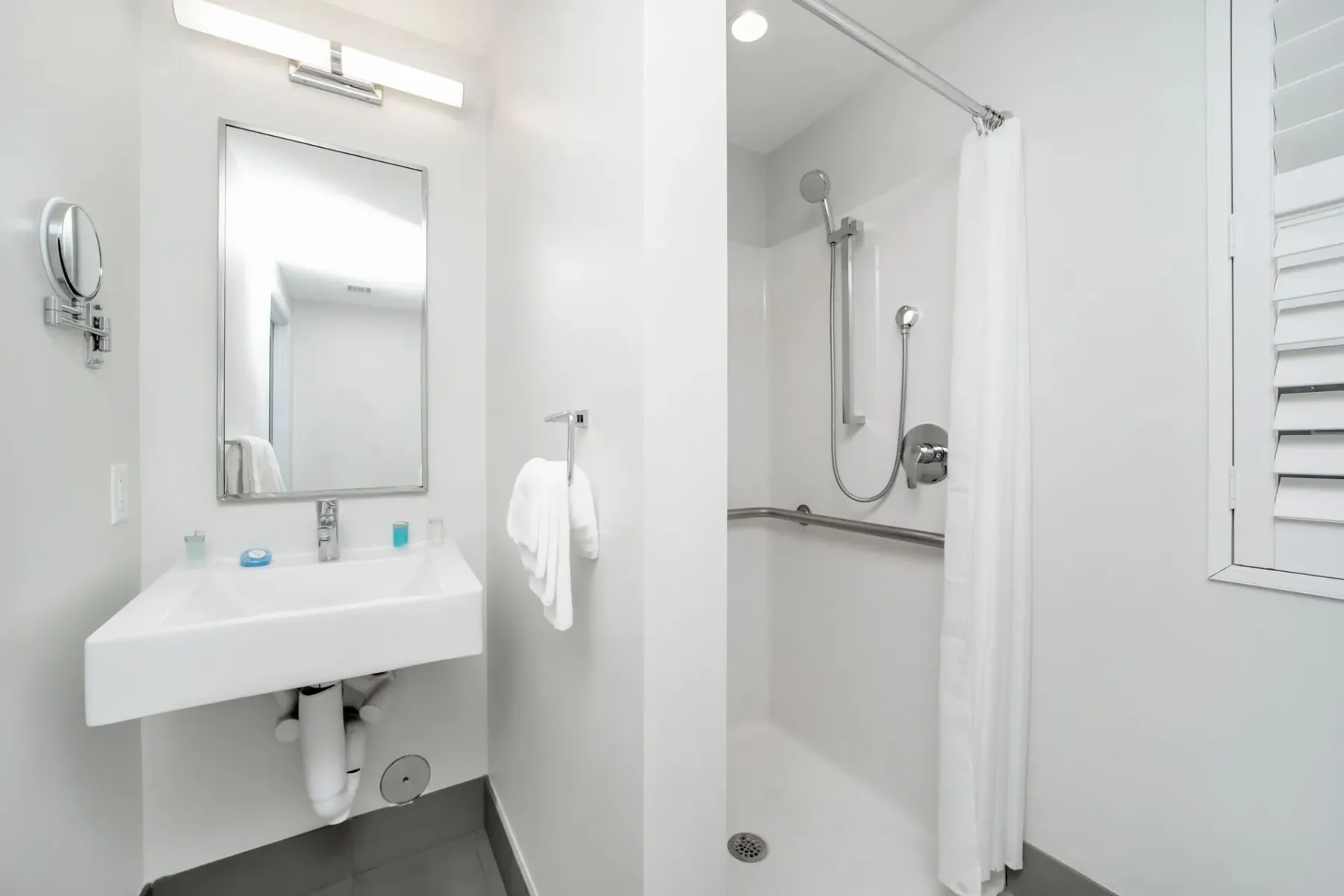 Petite bathroom with a sink, shower, and shuttered window.