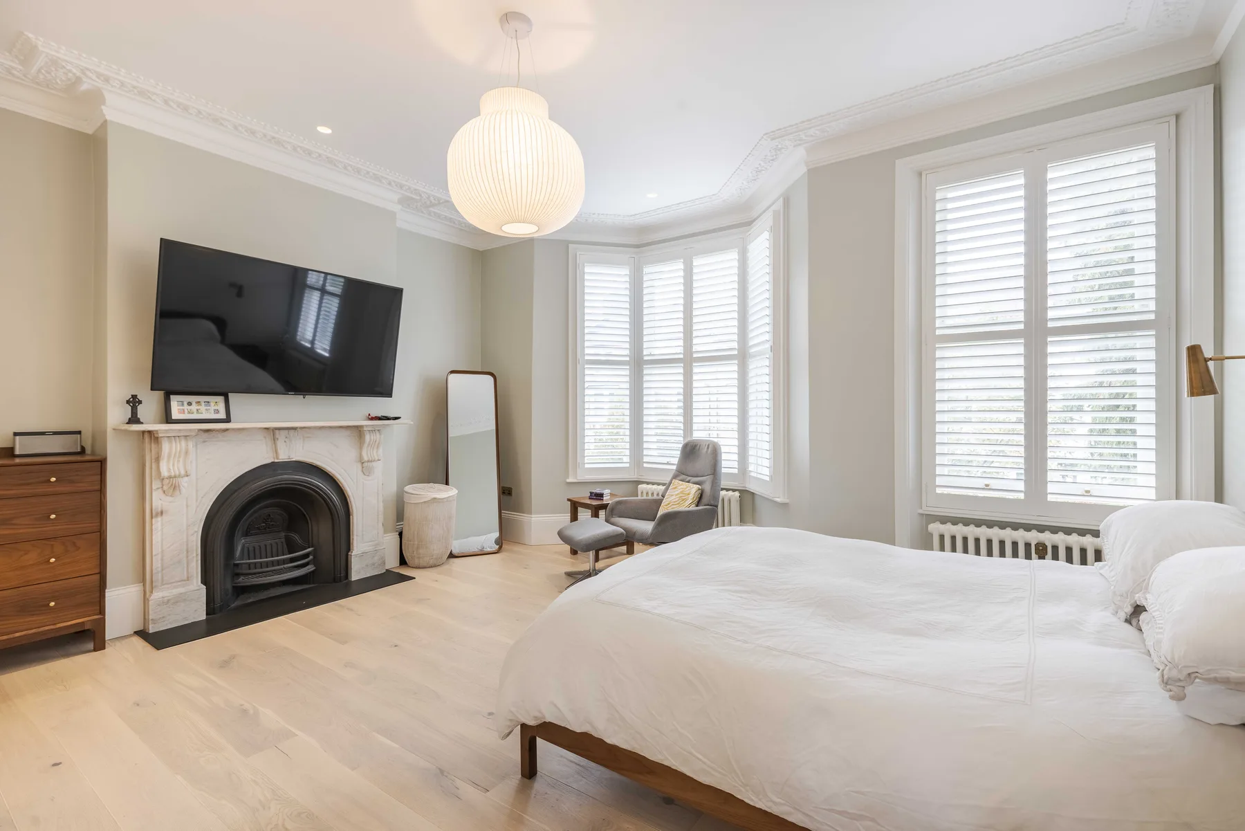 A bright bedroom with a large bed, a fireplace, and tall shuttered windows.