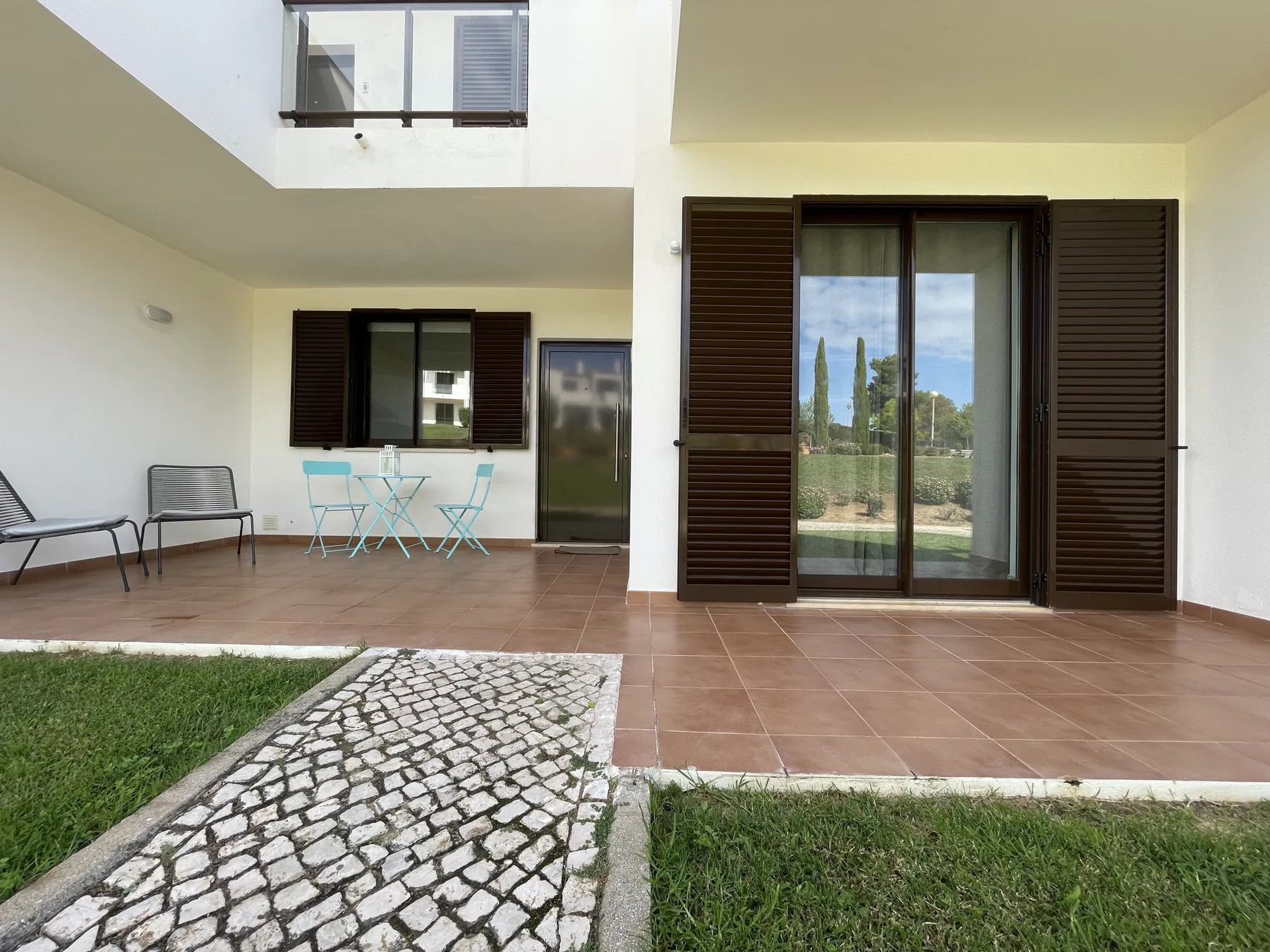 A ground-level patio with outdoor seating and dark shutters beside a cobbled path.
