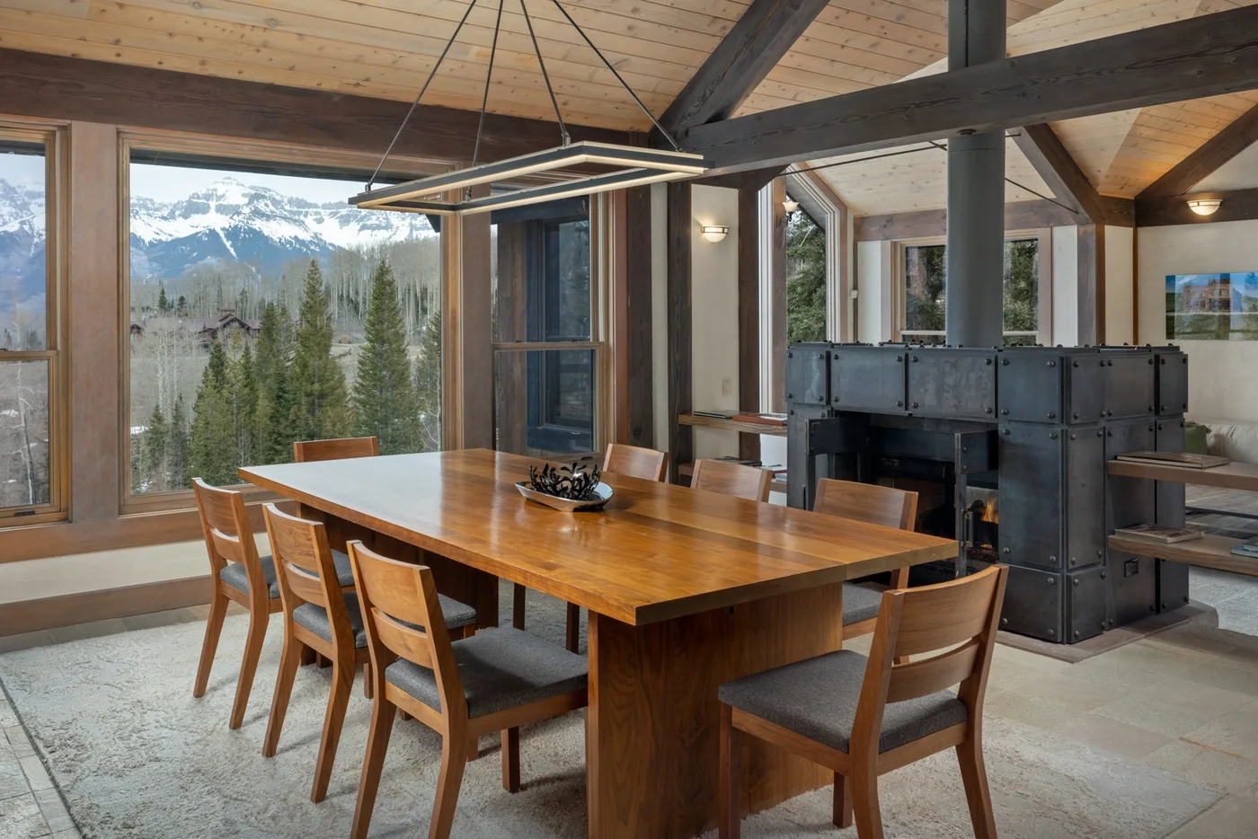 A dining area with a wooden table and chairs beside large windows overlooking a mountain landscape.