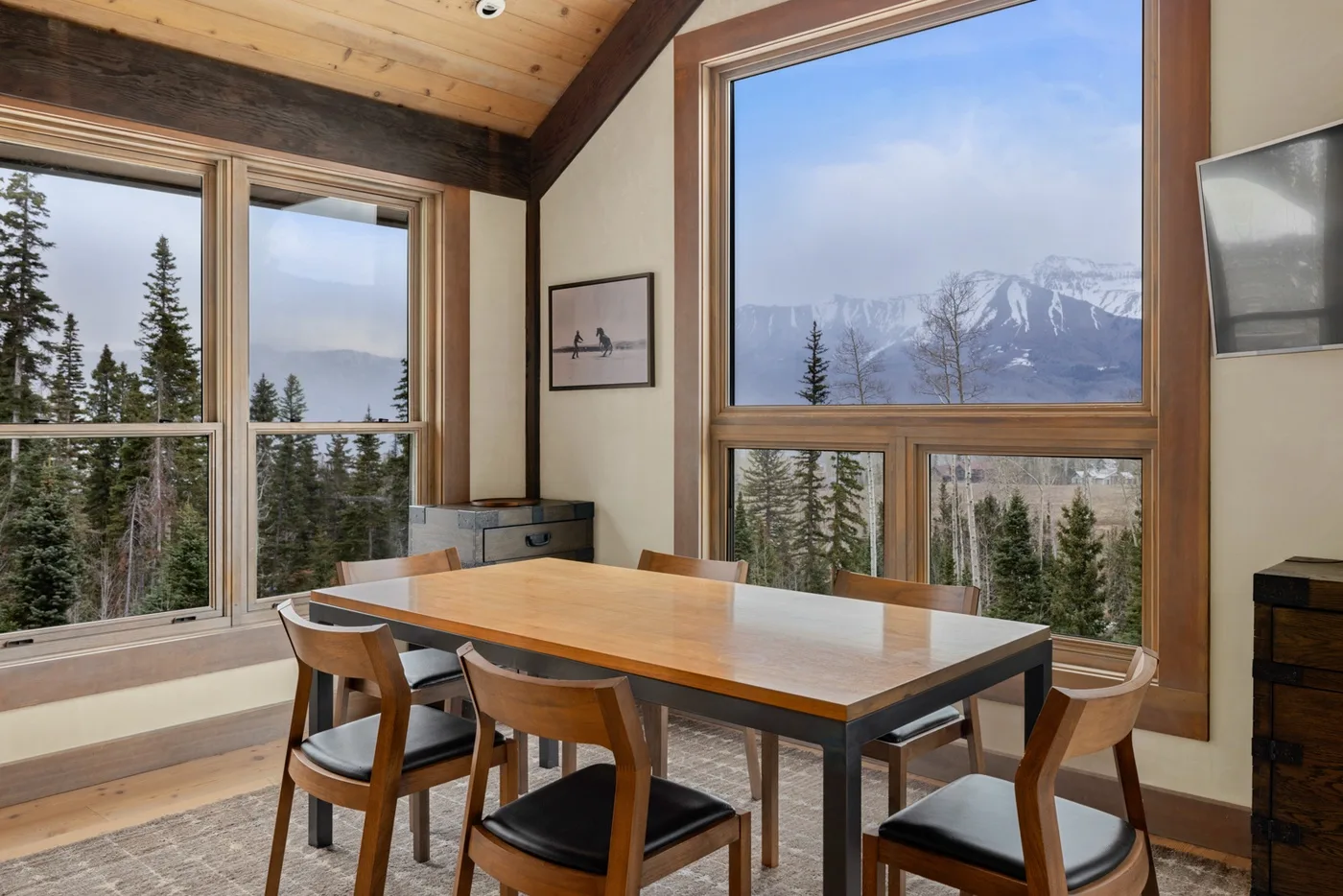 A dining area with a large table and chairs beside windows overlooking trees and mountains.