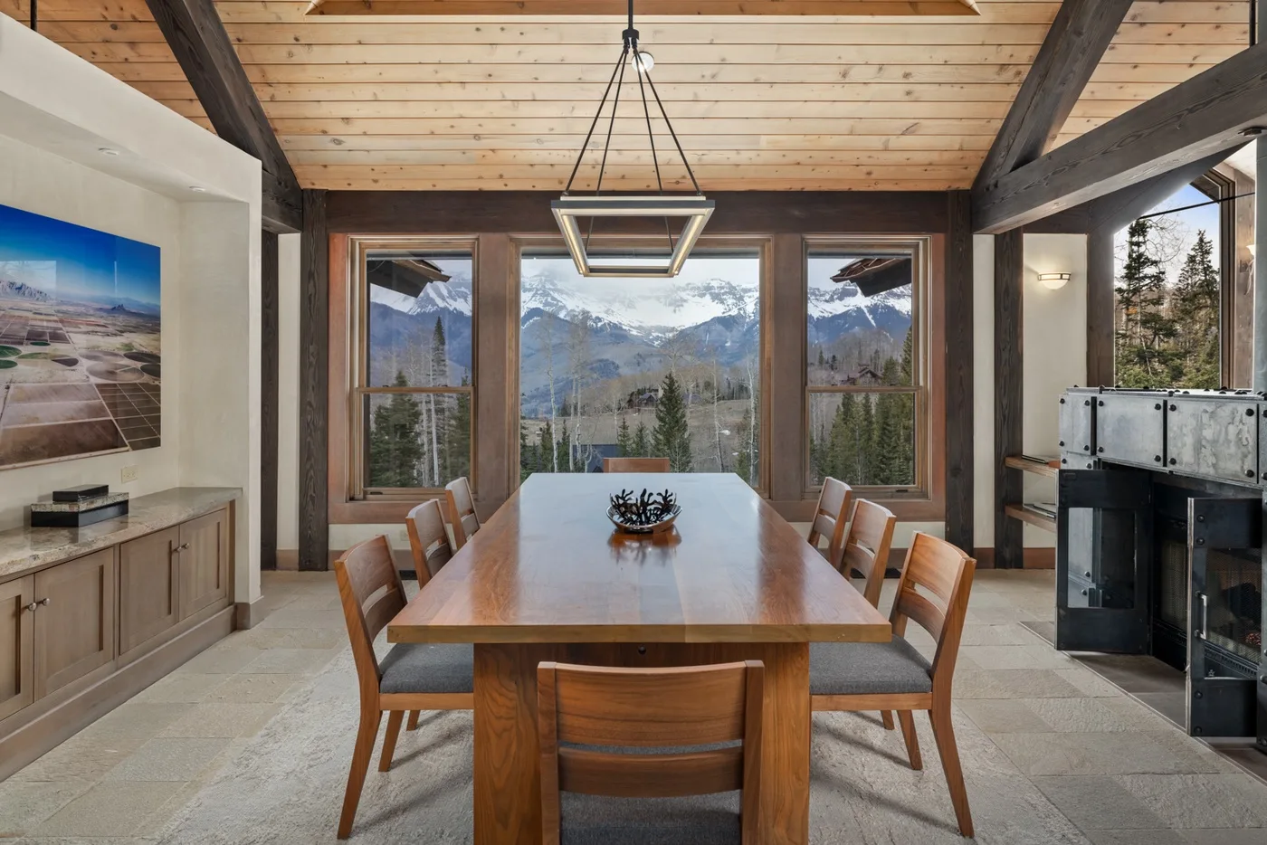 A dining area with a long wooden table and mountain views through large windows.