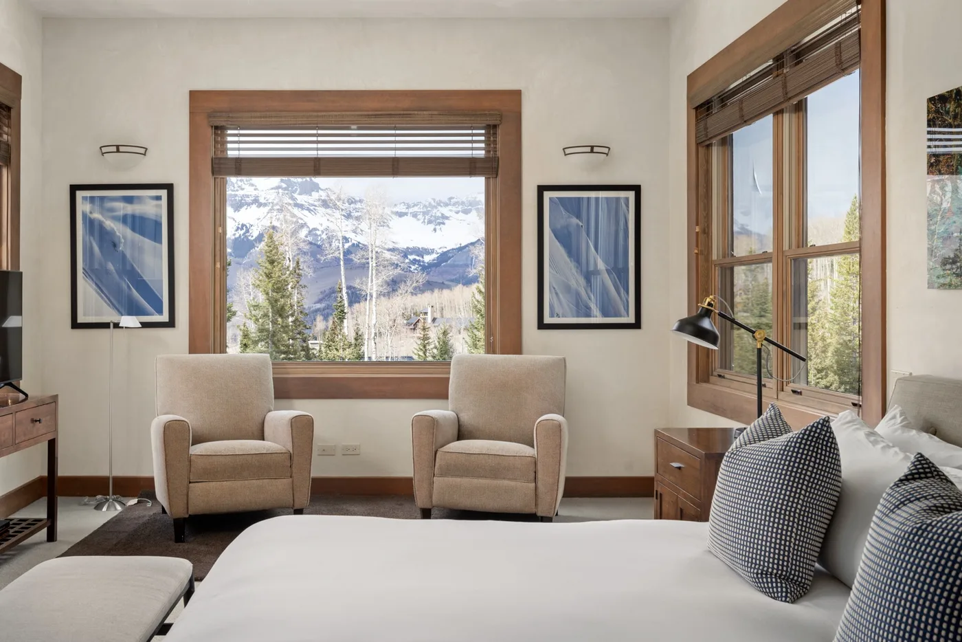 A bright bedroom has two upholstered seats beside windows overlooking a mountain landscape.