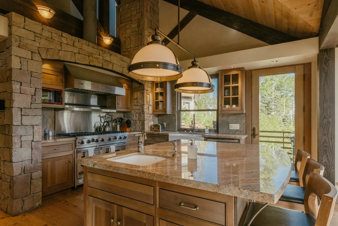 A spacious kitchen with a large island, stone walls, and windows overlooking trees.
