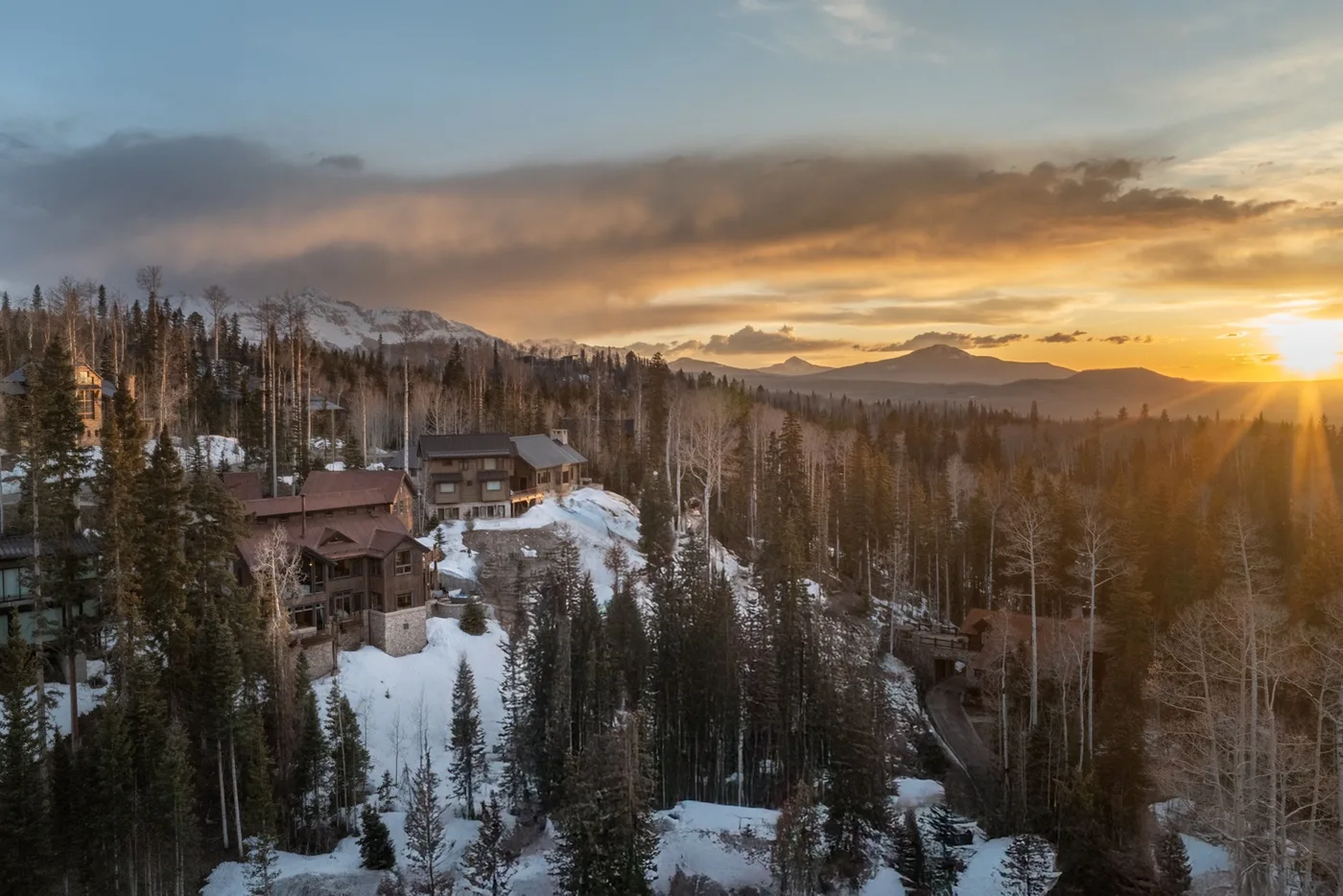 Snow-covered cabins sit among forested hills beneath a glowing sunset.