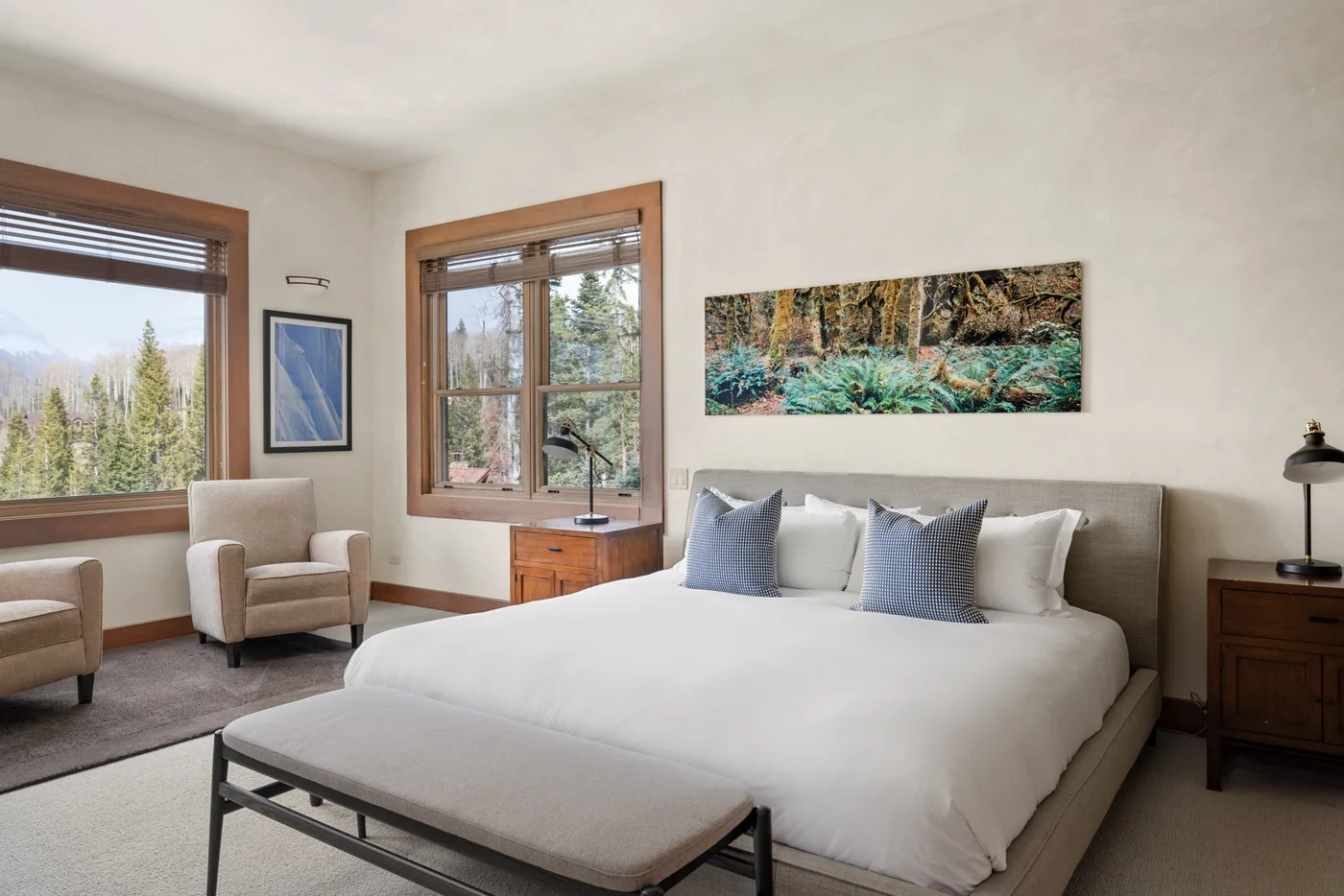 A bright bedroom with a large bed, a cushioned bench, and windows overlooking trees.