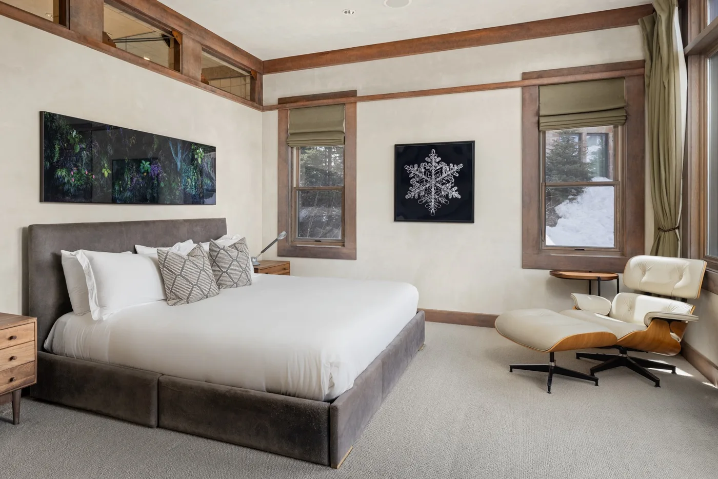 A bedroom with a large bed, wall art, and a lounge chair beside the windows.