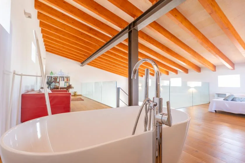 A bright loft bedroom features a freestanding bathtub beneath exposed wooden ceiling beams.