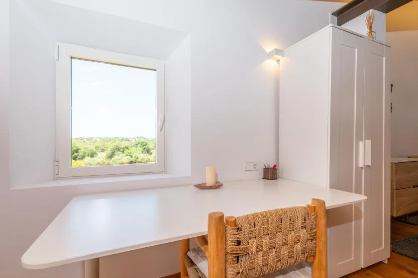 A bright workspace with a white desk, woven chair, and window overlooking greenery.
