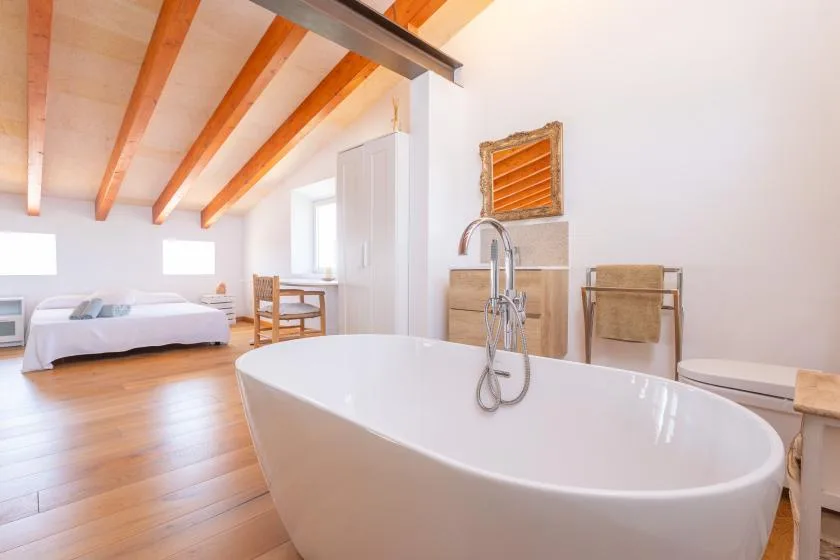 A bright bedroom and bathroom feature a freestanding bathtub beneath exposed wooden ceiling beams.