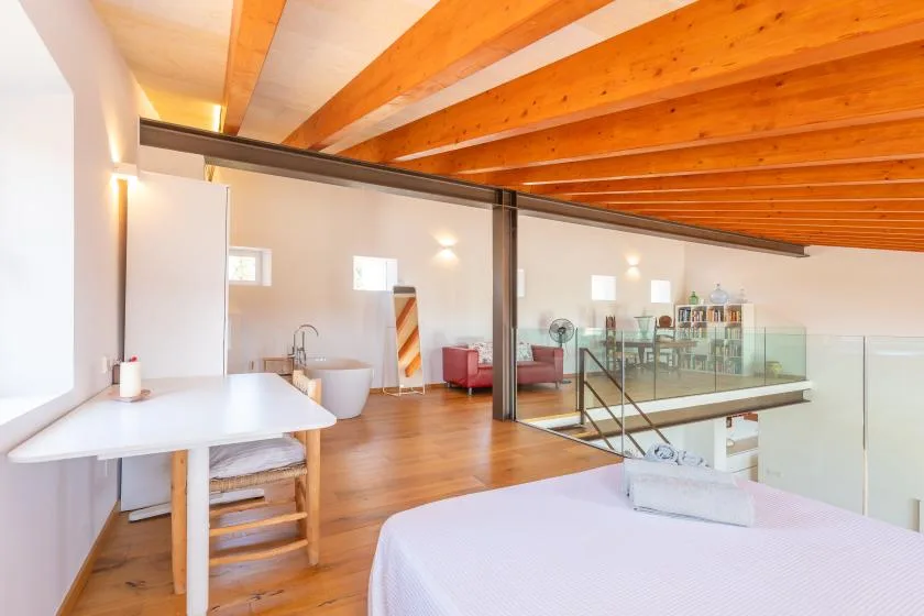 A bright loft bedroom opens onto a glass-railed mezzanine beneath exposed wooden beams.