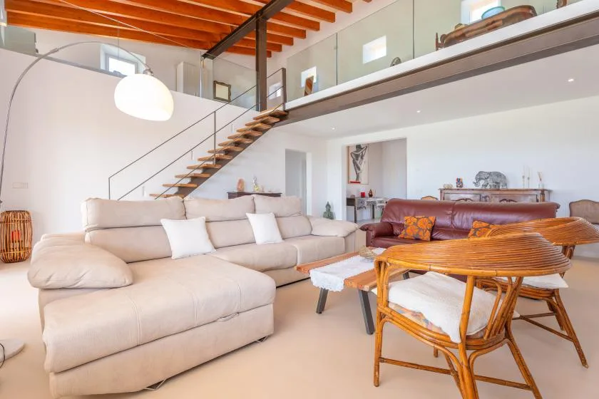 A bright living room with a large sectional, a dining table, and a mezzanine staircase.