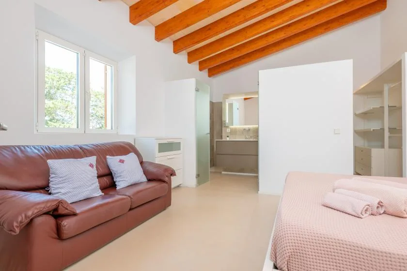 A bright bedroom with a bed, a brown sofa, and exposed wooden ceiling beams.