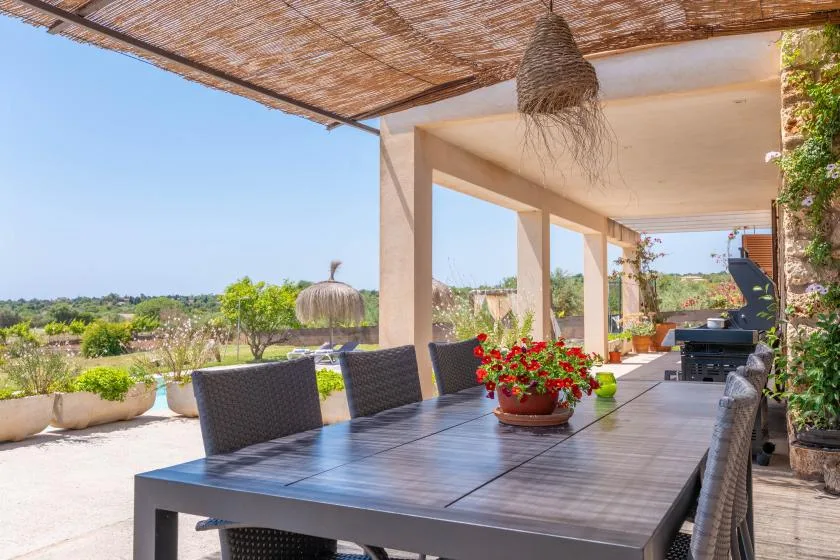 A shaded outdoor dining area with a long table and potted flowers overlooks greenery.