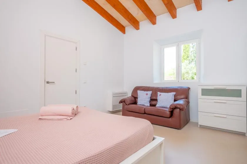 A bright bedroom with a bed, a brown loveseat, and a window.