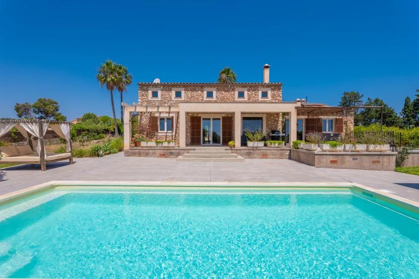 A modern house stands beyond a swimming pool, with a paved patio in front.