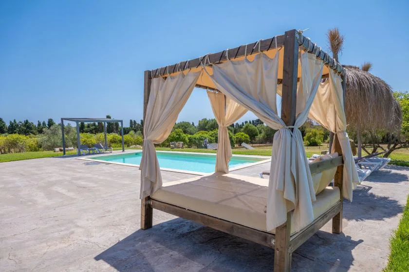 An outdoor cabana with draped curtains sits beside a swimming pool.
