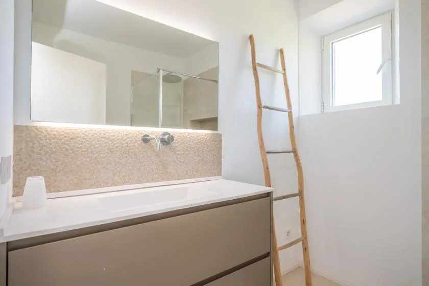 A bright bathroom with a wide vanity, wall mirror, and high window.