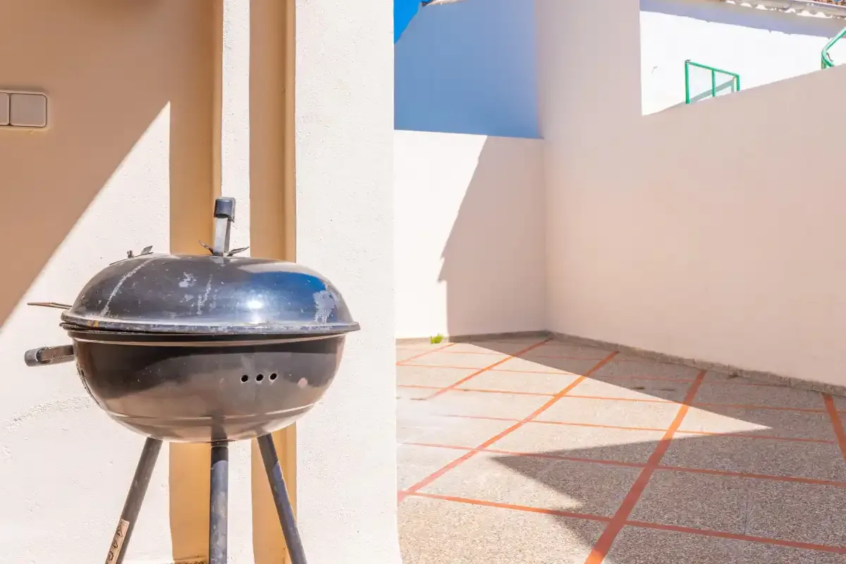 A sunlit outdoor patio with a charcoal grill beside a white wall.
