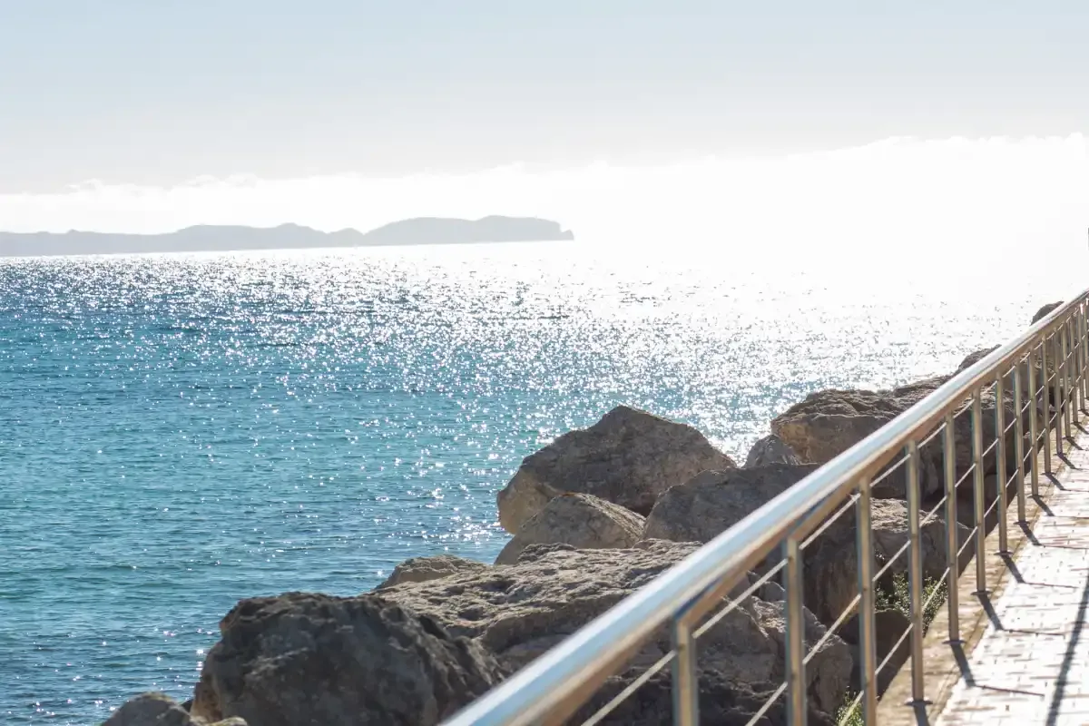 A seaside walkway with a metal railing runs beside rocky shoreline and blue water.