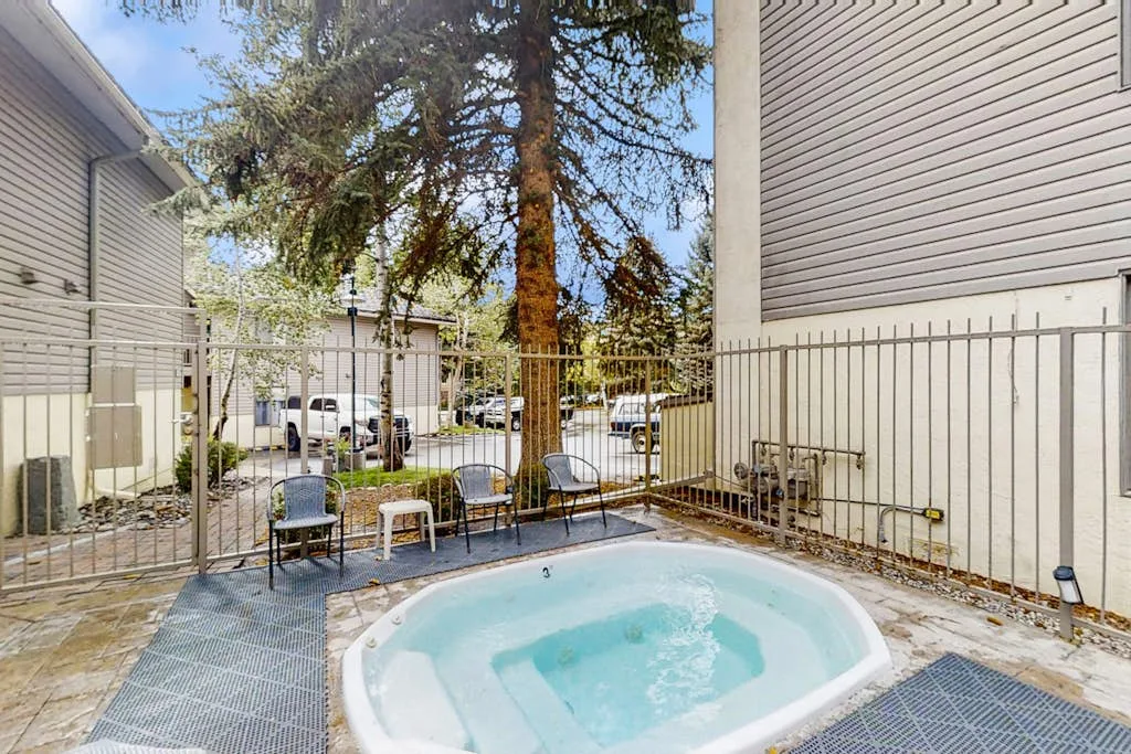 An outdoor hot tub sits on a paved patio between light-sided buildings and a metal fence.