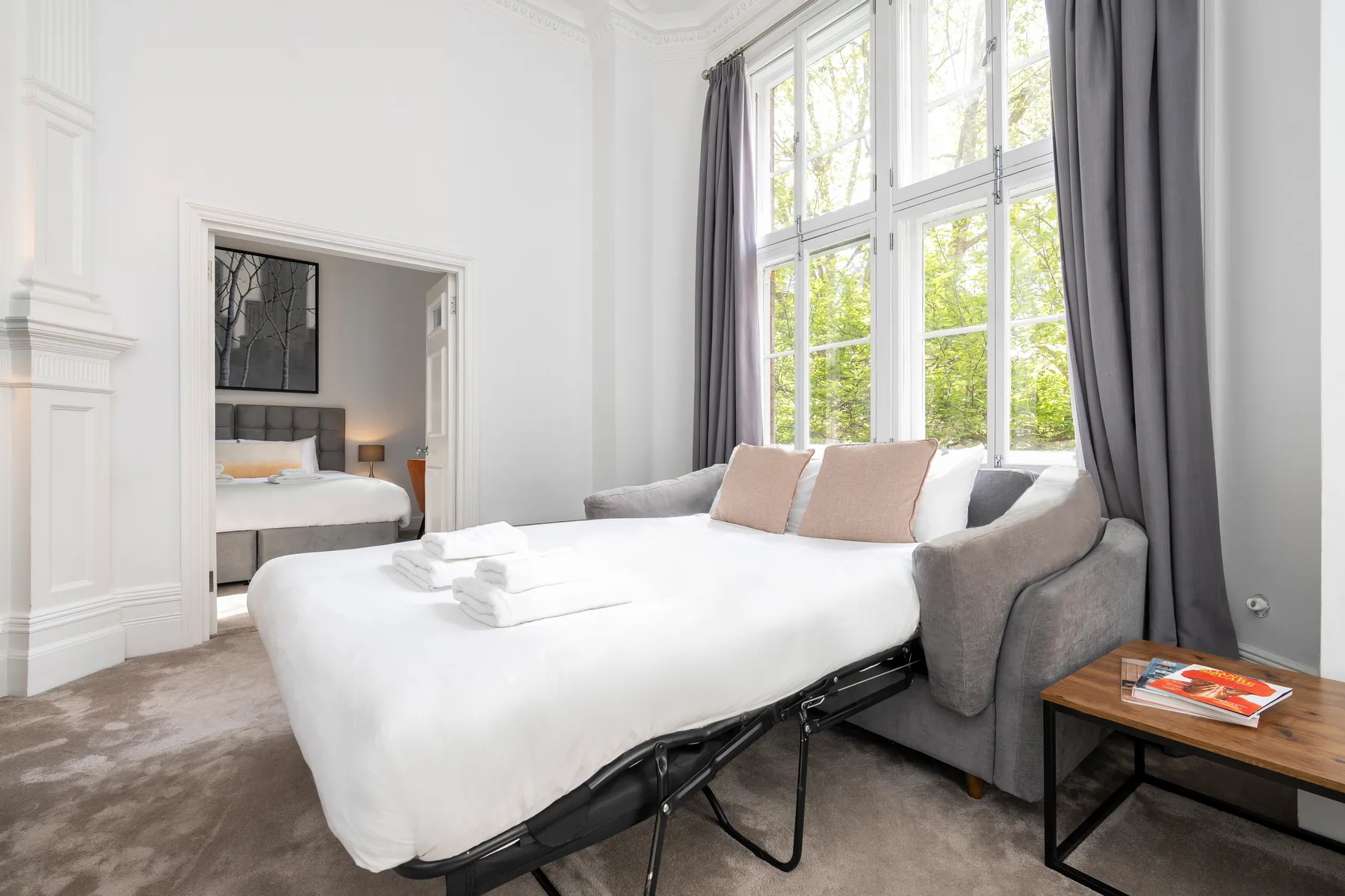 Un dormitorio luminoso con una cama junto a una gran ventana con cortinas grises.