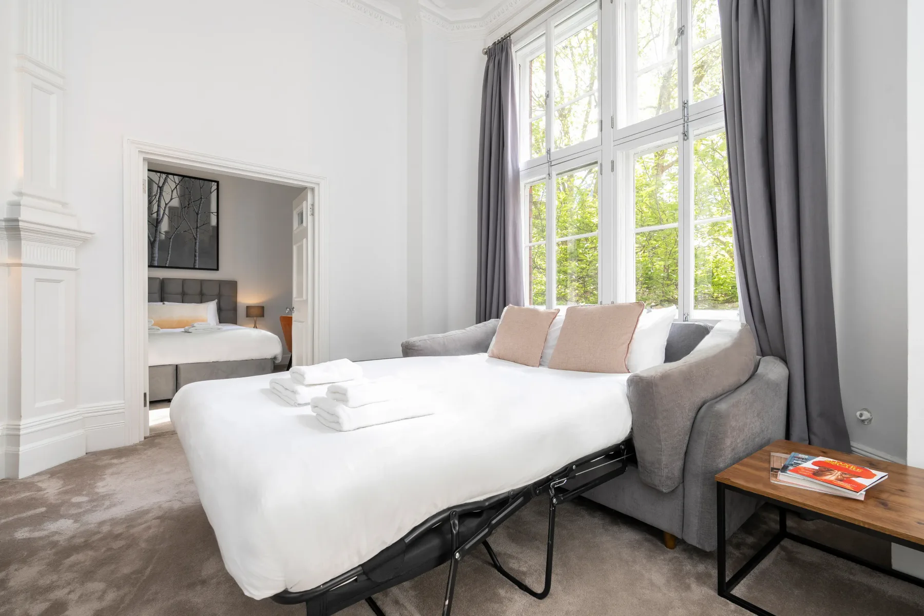 Un dormitorio luminoso con una cama blanca, asientos grises y ventanas altas.