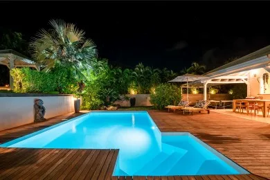 A brightly lit swimming pool sits beside a wooden deck and outdoor seating at night.