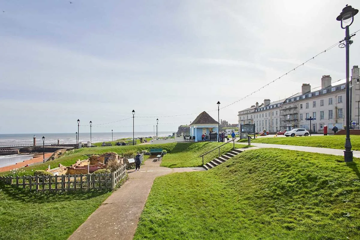 Un paseo junto al mar con un parque verde, un sendero y edificios frente al agua.